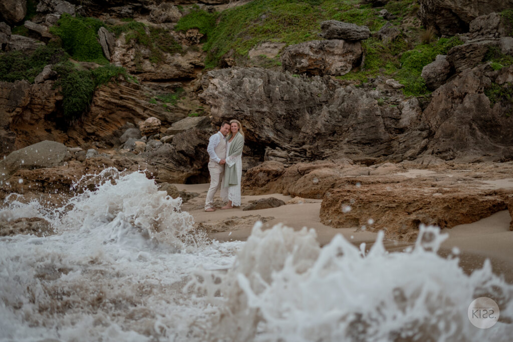 A couple standing on rocks as a wave breaks in the foreground