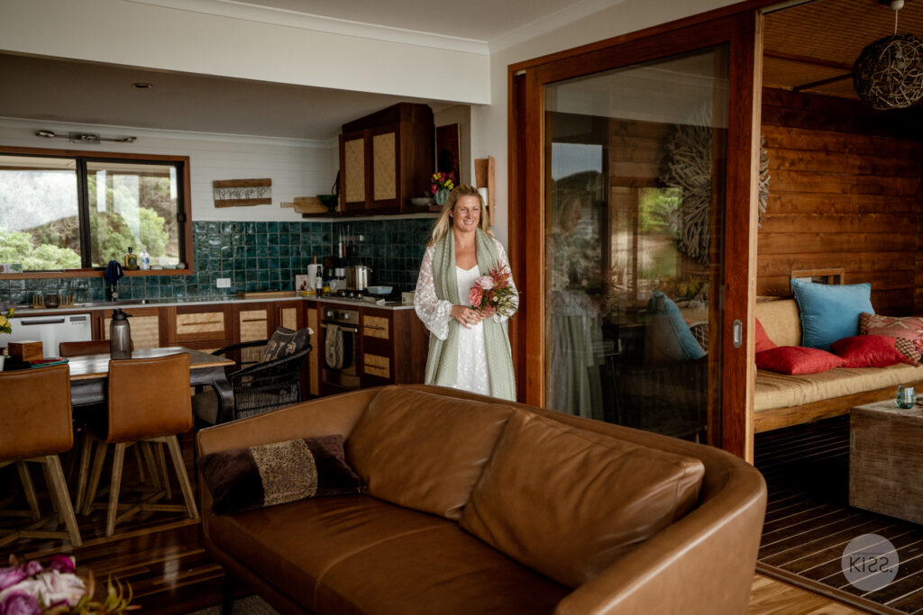 A bride standing inside a timber cottage overlooking the coast